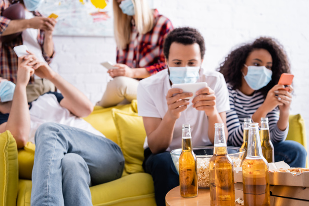 bottles of beer and bowl of popcorn near multiethnic friends in medical masks messaging on mobile phones on blurred background - Photo, image