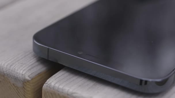 Κοντινό πλάνο ενός μέρους μιας μαύρης οθόνης ενός νέου smartphone σε ξύλινο πάγκο φόντο. Πάμε. Νέα απενεργοποιημένη συσκευή είναι αριστερά σε έναν πάγκο. - Πλάνα, βίντεο