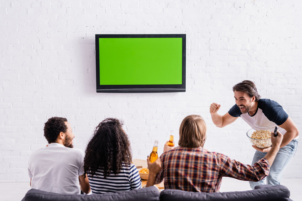 excited man holding popcorn and showing winner gesture near multiethnic friends watching championship - Фото, изображение