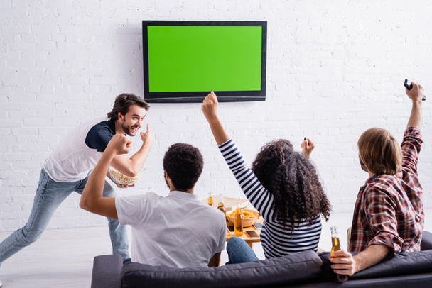 ενθουσιασμένος άνθρωπος που δείχνει σε LCD τηλεόραση στον τοίχο κοντά σε πολυπολιτισμικούς φίλους βλέποντας τον αθλητισμό ανταγωνισμού - Φωτογραφία, εικόνα