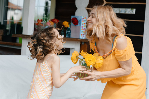 Mom and daughter in dresses are sitting on the terrace near the flowers hugging each other in the background flowers. Summer holidays. Nature and fresh air. Landscape and design. - Foto, Imagem