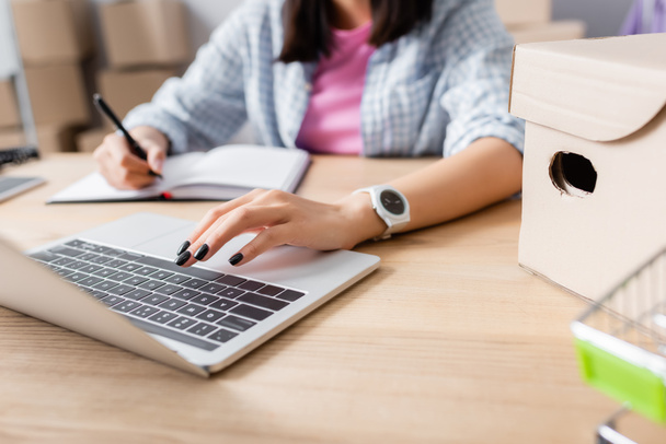 Widok kobiety korzystającej z laptopa podczas pisania w notebooku przy biurku z pudełkiem kartonowym na rozmytym tle - Zdjęcie, obraz