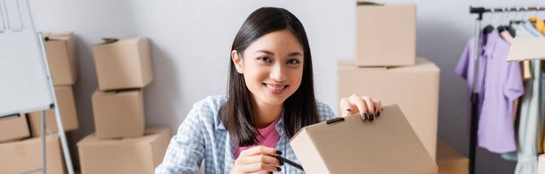 Happy asian volunteer looking at camera while opening carton box in charity center on blurred background, banner - Photo, Image