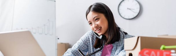 Sonriendo asin voluntario hablando por teléfono y mirando a la computadora portátil en el centro de caridad en primer plano borrosa, pancarta - Foto, Imagen