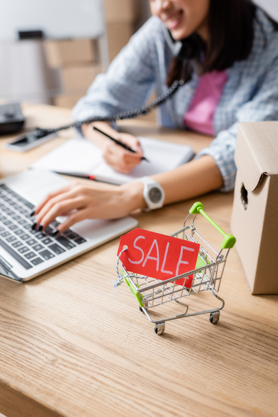 Close up view of price tag with sale lettering in shopping cart near carton box on desk with blurred woman on background - Photo, Image