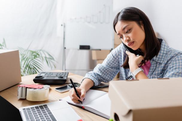 Pensativo asiático voluntario hablando por teléfono mientras escribe en portátil en el escritorio con cajas de cartón en primer plano borrosa - Foto, Imagen