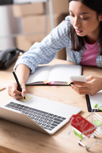 Volontario asiatico con scrittura smartphone su adesivo mentre seduto alla scrivania con notebook su sfondo sfocato - Foto, immagini