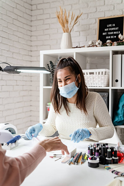 Manicure and pedicure salon, covid-19 and social distance. Manicure master in rubber gloves and mask talking with the client in beauty studio interior - Fotó, kép