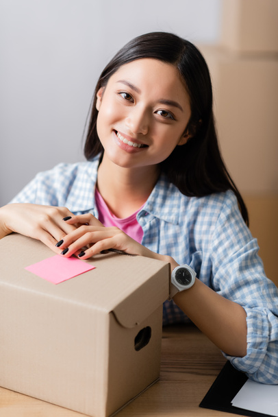 Hymyilevä aasialainen nainen katselee kameraa lähellä laatikkoa tahmea viesti hyväntekeväisyyskeskuksessa  - Valokuva, kuva