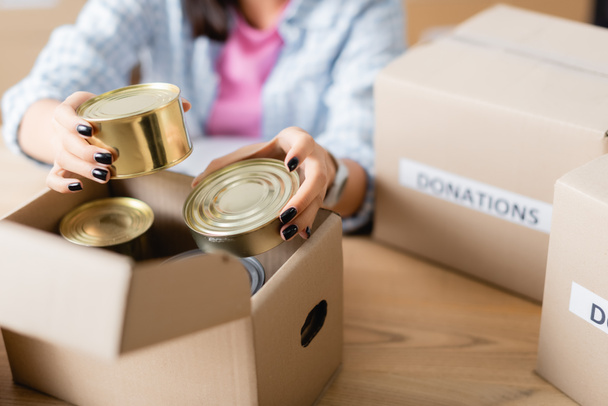 Cropped view of woman holding canned food near carton box in charity center on blurred background  - Photo, image