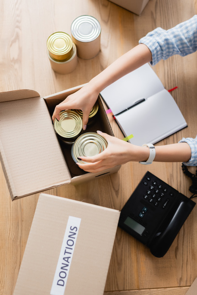 Κάτοψη της εθελοντικής συσκευασίας κονσερβοποιημένων τροφίμων σε κουτί κοντά στο σημειωματάριο και τηλέφωνο στο τραπέζι  - Φωτογραφία, εικόνα