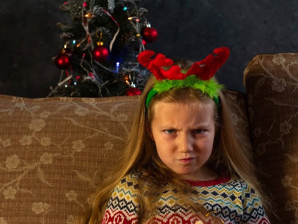 Vakava pieni tyttö punaisissa peuran sarvissa irvistelee katsellen kameran edessä kotikuvaa. Loukkaantunut vihainen hampaaton lapsi uudenvuodenaattona joulukuusen valot. Perhelomien juhlan käsite. - Valokuva, kuva