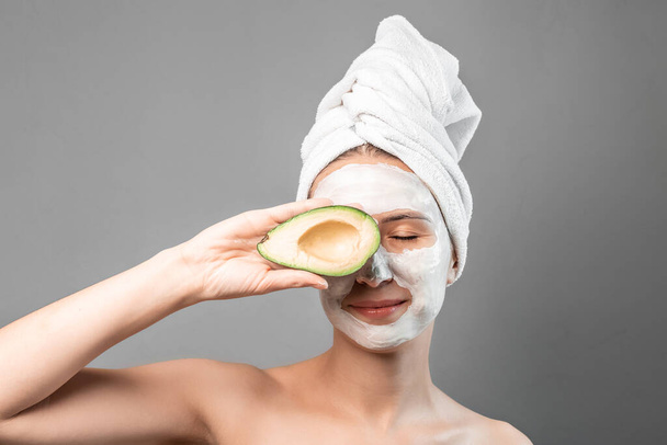 Beauty portret van lachende brunette vrouw in een handdoek op het hoofd met wit voedende masker of crème op het gezicht en verse avocado in de hand - Foto, afbeelding