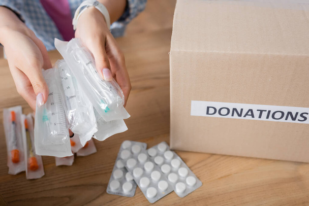 Vista ritagliata di siringhe nelle mani del volontario vicino a pillole e scatola con lettere di donazioni  - Foto, immagini