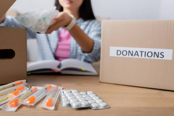 Pillen en spuiten op tafel bij vrijwilliger en dozen met donaties belettering op wazige achtergrond  - Foto, afbeelding