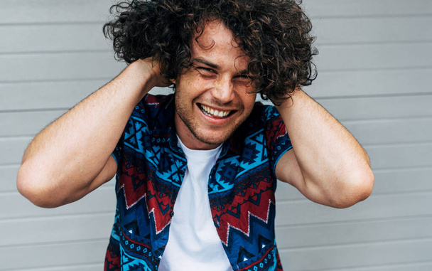 Cheerful male wearing casual trendy outfits enjoying the good day. A young man smiling broadly, looking at the camera, and arranges his curly hair on the city street while waiting for his friend. - Foto, Imagen