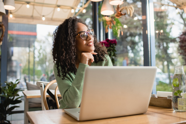 donna afro-americana sognante in occhiali sorridente mentre guarda lontano vicino al computer portatile sul tavolo  - Foto, immagini