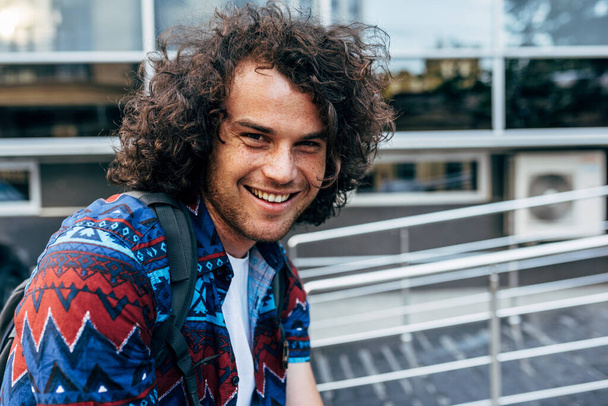 A male with curly hair wearing a casual trendy outfit sitting outside and looking at the camera. Smiling student man with a backpack waiting for his colleagues on the city street. - Fotografie, Obrázek