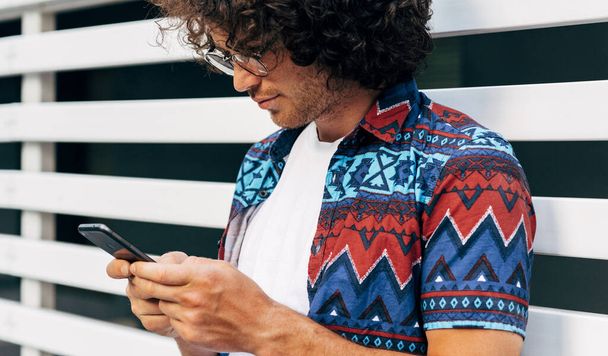 Closeup image of a young man checking his phone outside. Male with curly hair reading a message on a mobile phone in the city street. - 写真・画像