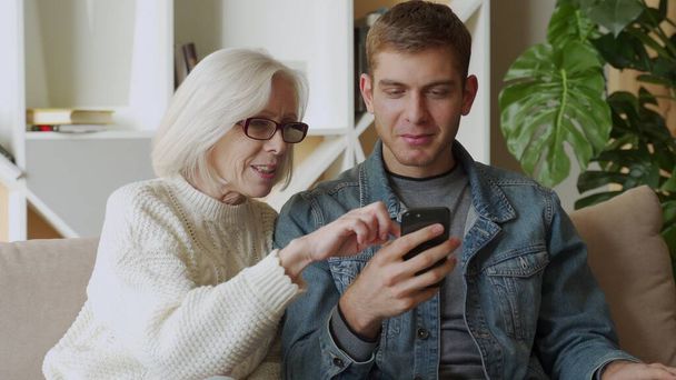 Smiling middle-aged mother rest with grown-up son using smartphone together, family weekend with senior mom browsing wireless Internet on cellphone - Фото, изображение