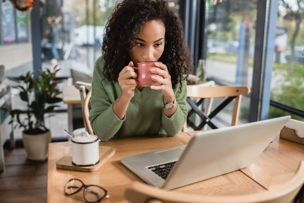 afro-amerikai nő iszik kávét csészéből, miközben nézi laptop  - Fotó, kép