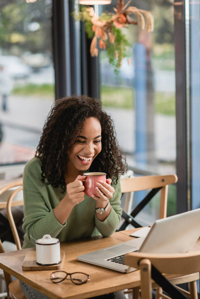 χαρούμενη αφρικάνικη αμερικανική γυναίκα κρατώντας κύπελλο, ενώ κοιτάζοντας φορητό υπολογιστή στο καφέ  - Φωτογραφία, εικόνα