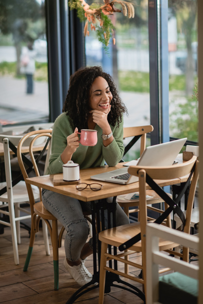 happy african american woman holding cup of coffee while looking at laptop in cafe - Foto, afbeelding