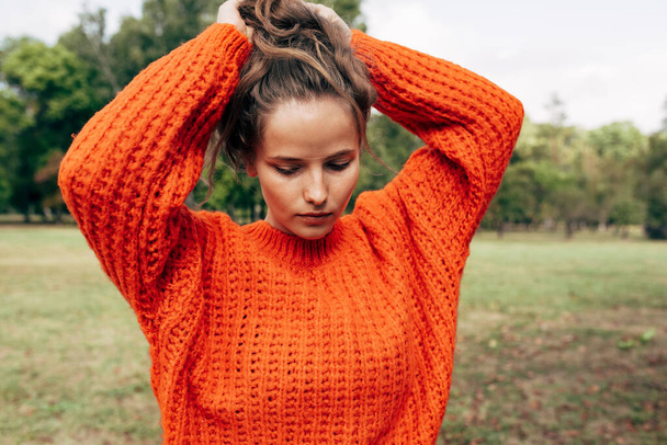 The young woman in an orange knitted sweater arranges her hair in a ponytail against a nature background. The gorgeous female has a serious expression, resting outdoors in the park. - Fotoğraf, Görsel