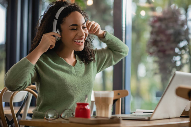 joyful african american woman listening podcast near laptop in cafe  - Fotografie, Obrázek