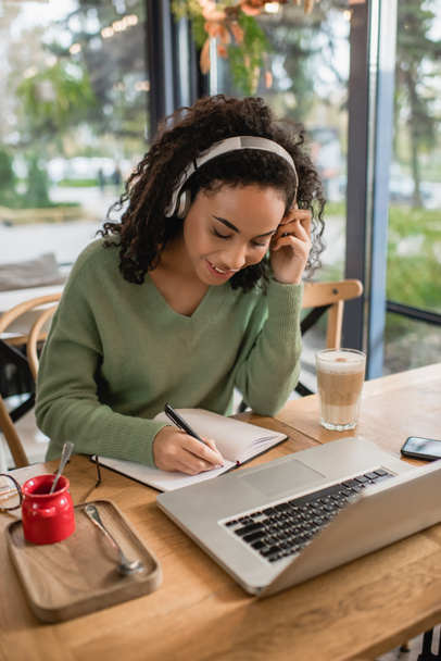 felice donna afroamericana ascolto podcast in cuffia e scrittura in notebook vicino laptop in caffè  - Foto, immagini
