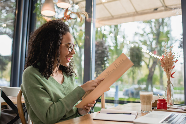 femme afro-américaine gaie dans les lunettes en regardant le menu près de l'ordinateur portable dans le café  - Photo, image