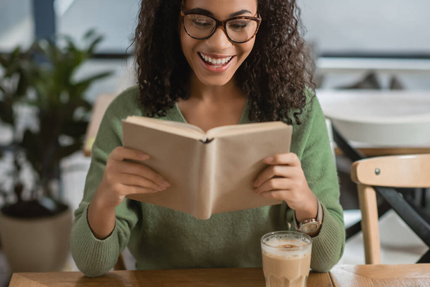 cheerful african american woman reading book near glass with latte in cafe - Foto, imagen