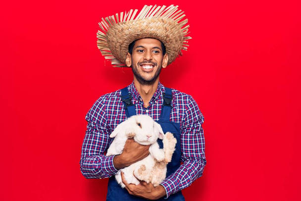 Fiatal latin farmer férfi kötényben és kalapban boldogan mosolyog. Álló mosollyal az arcán tartja imádnivaló nyúl felett elszigetelt piros háttér - Fotó, kép