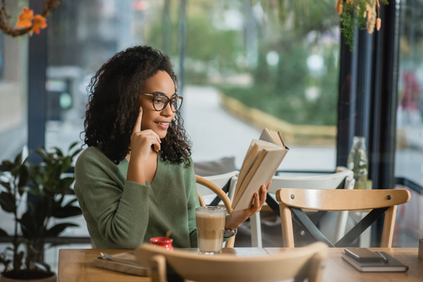 cheerful african american woman in eyeglasses reading book near glass with latte in cafe - Foto, Imagem