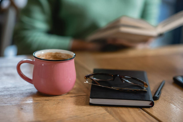 mug with coffee near eyeglasses, notebook and african american woman on blurred background  - Foto, Imagem