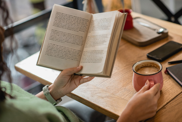 Teilansicht einer afrikanisch-amerikanischen Frau mit Buch und Tasse Cappuccino im Café - Foto, Bild