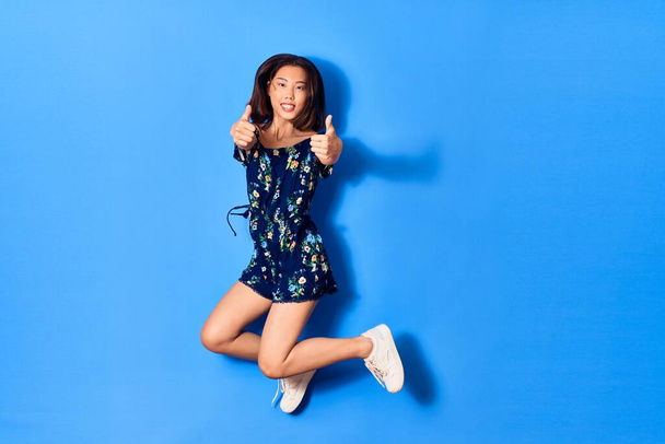 Young beautiful chinese woman wearing casual clothes smiling happy. Jumping with smile on face doing ok sign with thumbs up over isolated blue background - Photo, Image