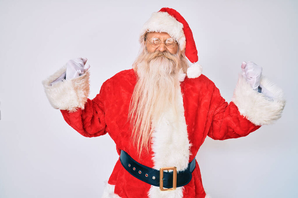 Staruszek z siwymi włosami i długą brodą w kostiumie Świętego Mikołaja bardzo szczęśliwy i podekscytowany wykonując gest zwycięzcy z podniesionymi ramionami, uśmiechnięty i wołający o sukces. koncepcja uroczystości.  - Zdjęcie, obraz