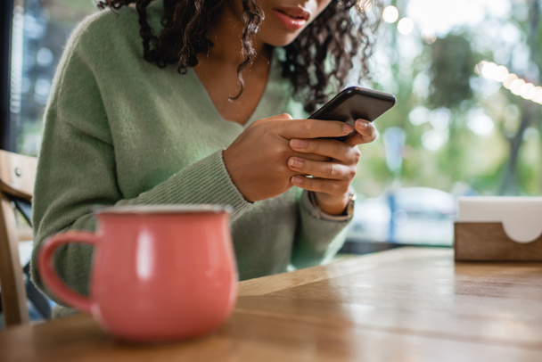 cropped view of african american woman texting on smartphone near cup on blurred foreground  - Фото, изображение