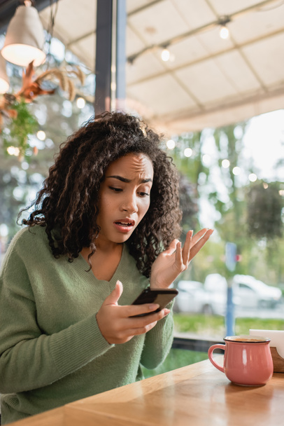 dissatisfied african american woman looking at smartphone while gesturing in cafe - Photo, image