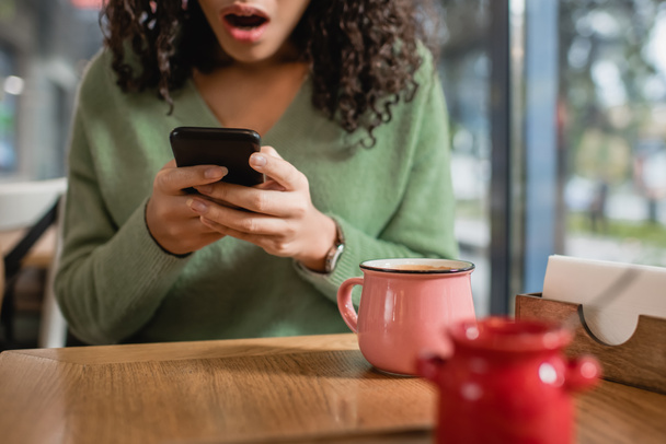 cup of coffee near shocked african american woman texting on smartphone on blurred foreground  - Photo, Image
