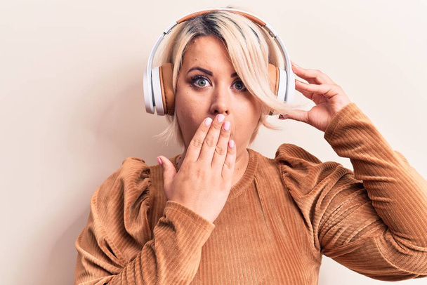 Mooie blonde plus size vrouw luisteren naar muziek met behulp van een koptelefoon over witte achtergrond bedekken mond met de hand, geschokt en bang voor fouten. Verrassende uitdrukking - Foto, afbeelding