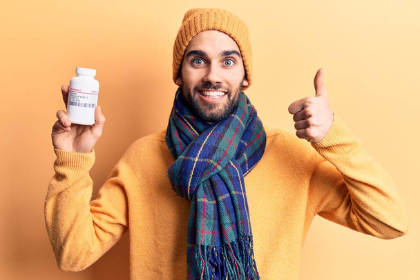 Young handsome man with beard holding bottle of pills smiling happy and positive, thumb up doing excellent and approval sign  - Photo, Image