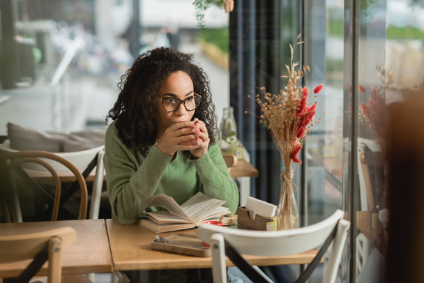 african american woman in eyeglasses drinking coffee near book on table in cafe - Photo, Image