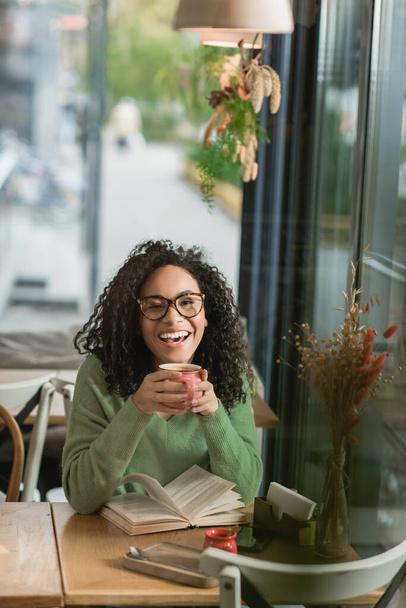 amazed african american woman in eyeglasses holding cup of coffee near book on table in cafe - Photo, Image