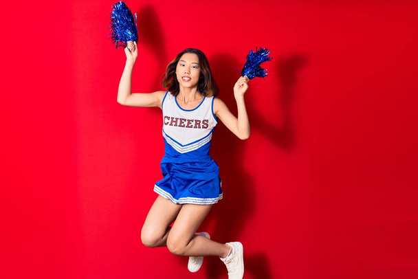 Young beautiful chinese girl smiling happy wearing cheerleader uniform. Jumping with smile on face using pompom over isolated red background - Photo, Image