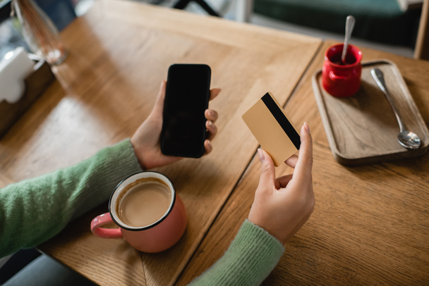 oříznutý pohled na africkou americkou ženu držící kreditní kartu v blízkosti smartphonu s prázdnou obrazovkou a pohár na stole - Fotografie, Obrázek
