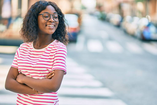 jong Afrikaans amerikaans meisje met armen gekruist lachend gelukkig staan in de stad. - Foto, afbeelding