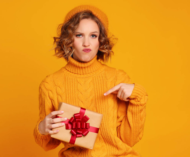 Unzufriedene junge Frau im Strickpulli am Hut schmollende Lippen und zeigt auf verpacktes Geschenk während der Weihnachtsfeier vor gelbem Hintergrund - Foto, Bild