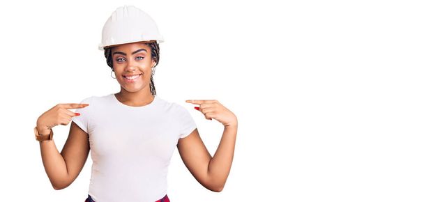 若いアフリカ系アメリカ人の女性は、硬い帽子をかぶって顔に笑顔で自信を持って服を構築し、指を誇りに思って幸せな自分自身を指しています.  - 写真・画像